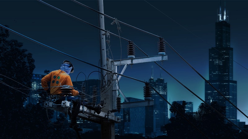 A utility line worker wearing a shirt with the Profisee logo on the back works on a power line to restore power to the city, which can be seen lighting up in the distance.